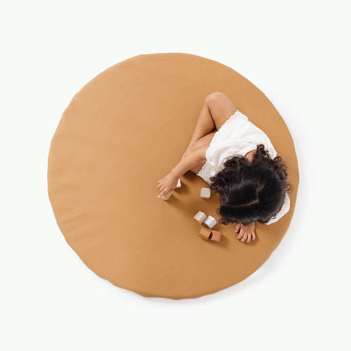 Overhead view of a girl lying on the round camel vegan leather padded play mat with wooden blocks