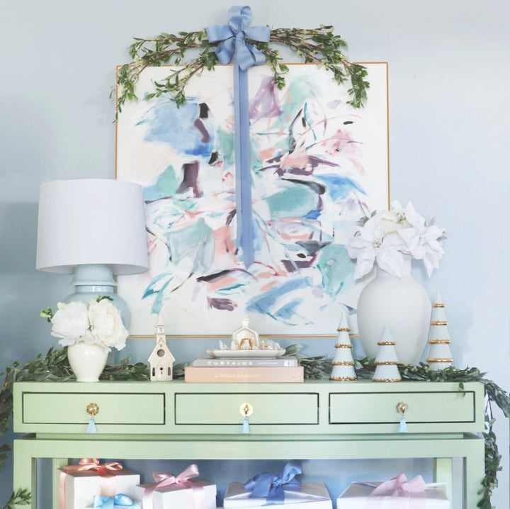 Mint console table styled with a ceramic village, blue lamp, garland, and abstract art for a pastel holiday entryway