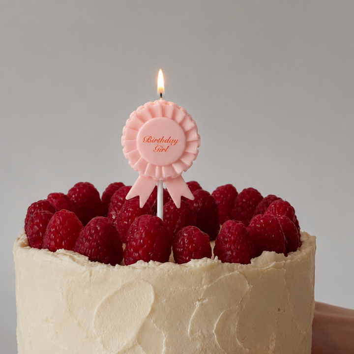 A close-up of a frosted cake topped with fresh raspberries and a pink ribbon-shaped candle that reads 'Birthday Love', with a hand holding the cake against a plain light gray background.