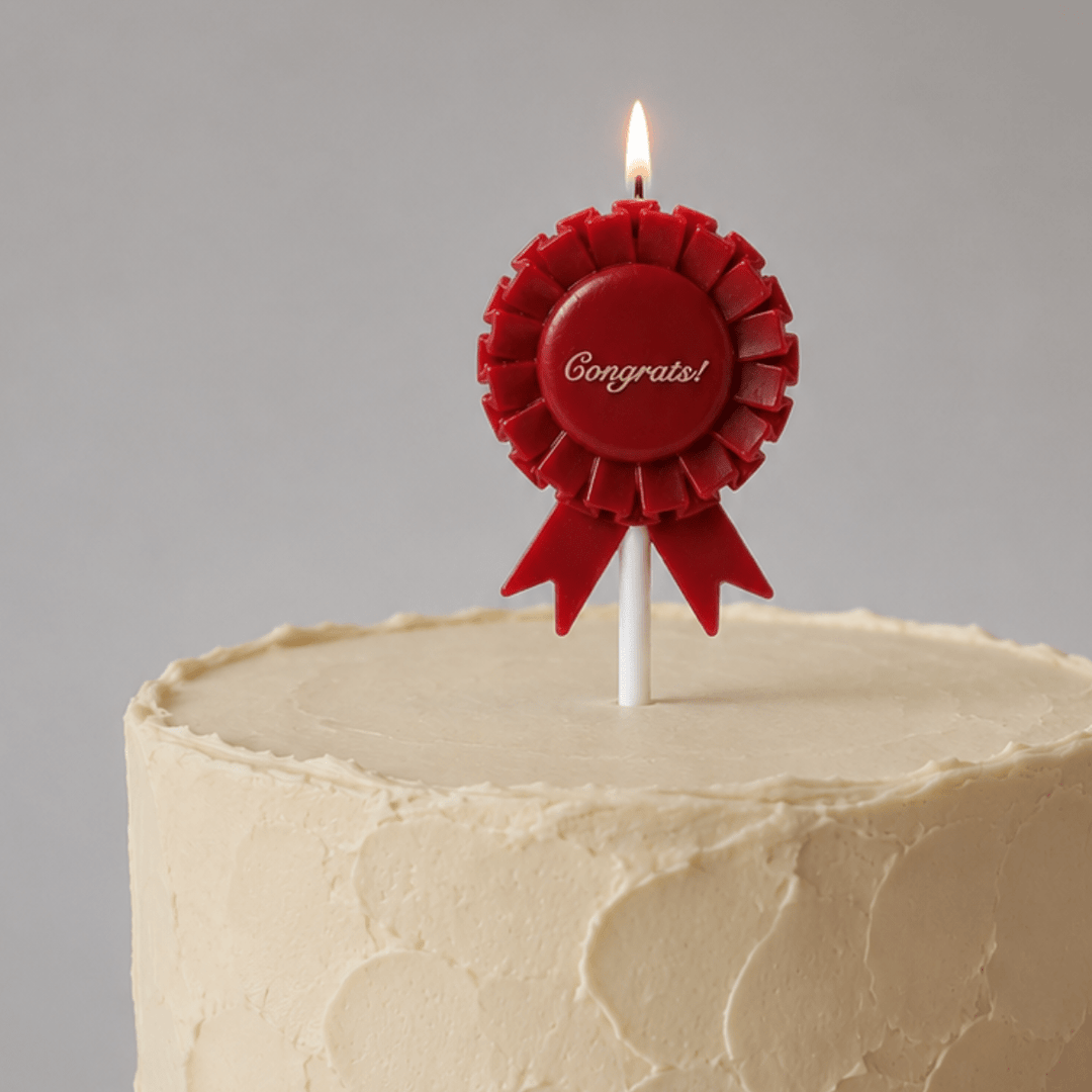 A red rosette-shaped birthday candle with a lit flame, printed with the word 'Congrats!' in white script, inserted into a cream-colored frosted cake against a neutral gray background.