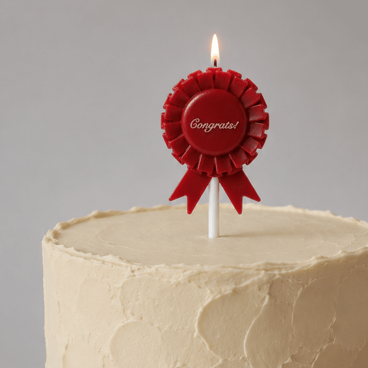 A red rosette-shaped birthday candle with a lit flame, printed with the word 'Congrats!' in white script, inserted into a cream-colored frosted cake against a neutral gray background.