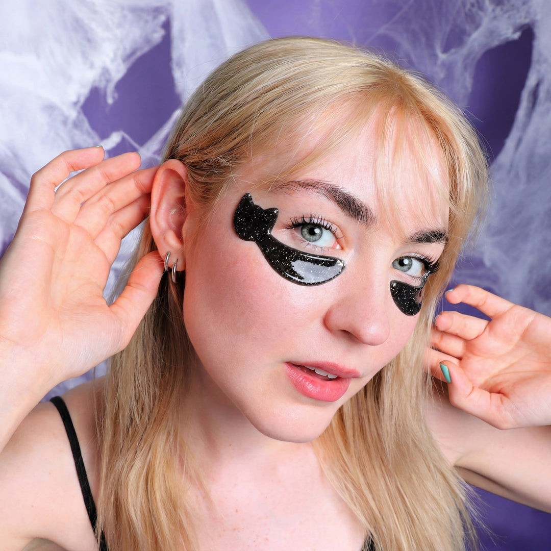 Model wearing sparkly black cat under eye patches against purple cobwebs, playful Halloween party prep
