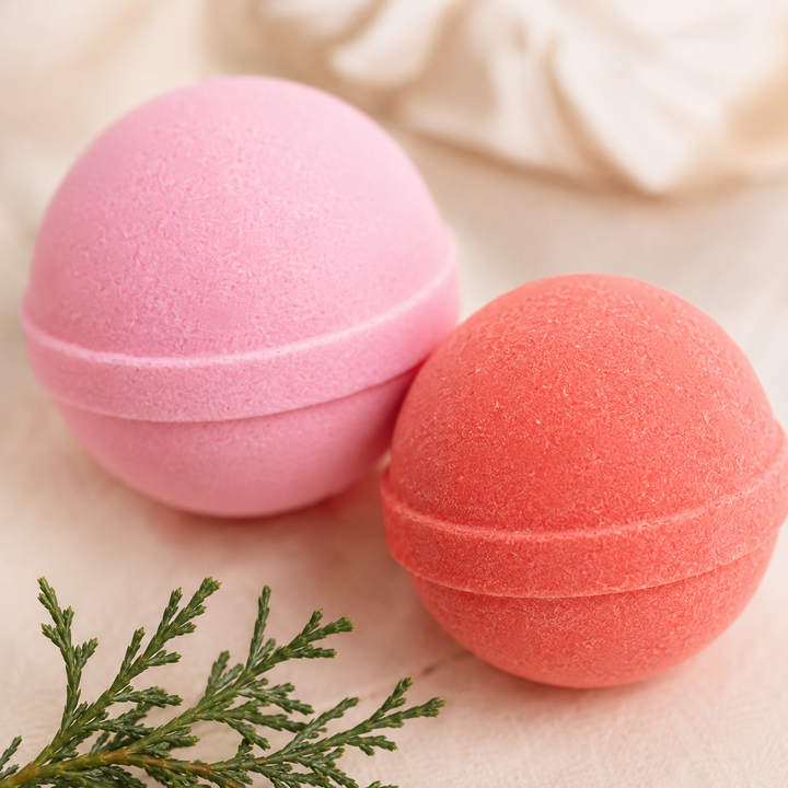Close-up of pink and coral mini bath bombs on linen with cedar sprigs, a cozy holiday bath moment