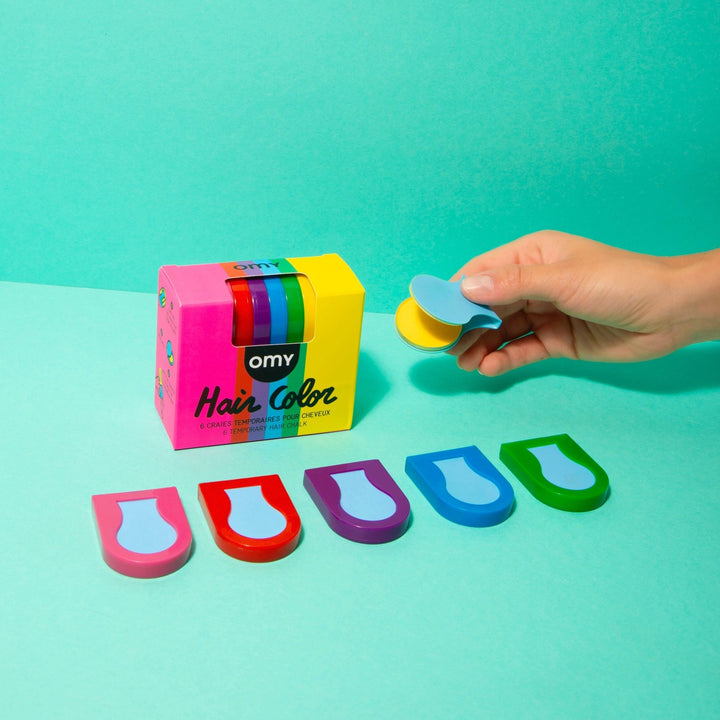 Rainbow hair chalk sticks with colorful chalk box and a hand holding a blue chalk piece on a teal background