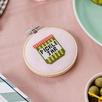 Finished pickle jar cross stitch hoop styled on a pink table runner with olives