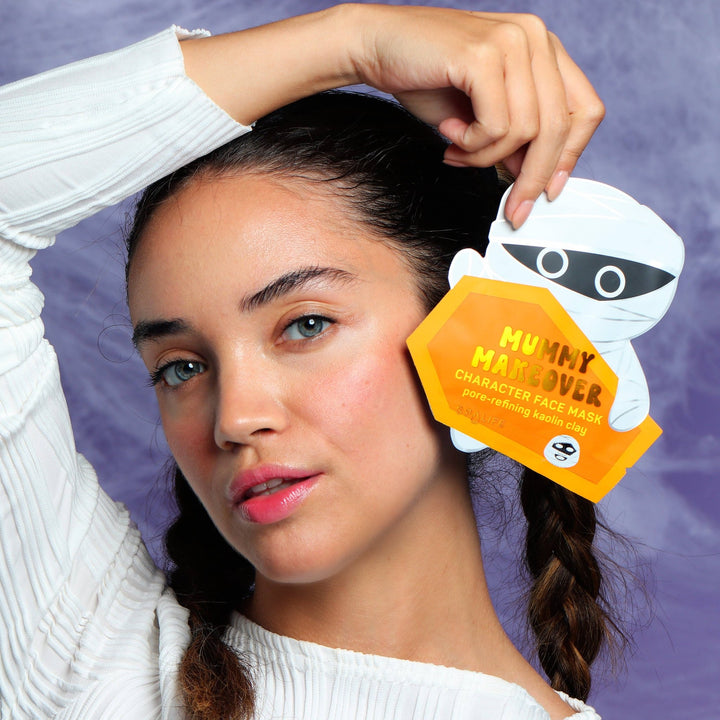 Model holding the mummy face mask pouch beside her cheek on a purple cobweb backdrop, a cute boo basket idea