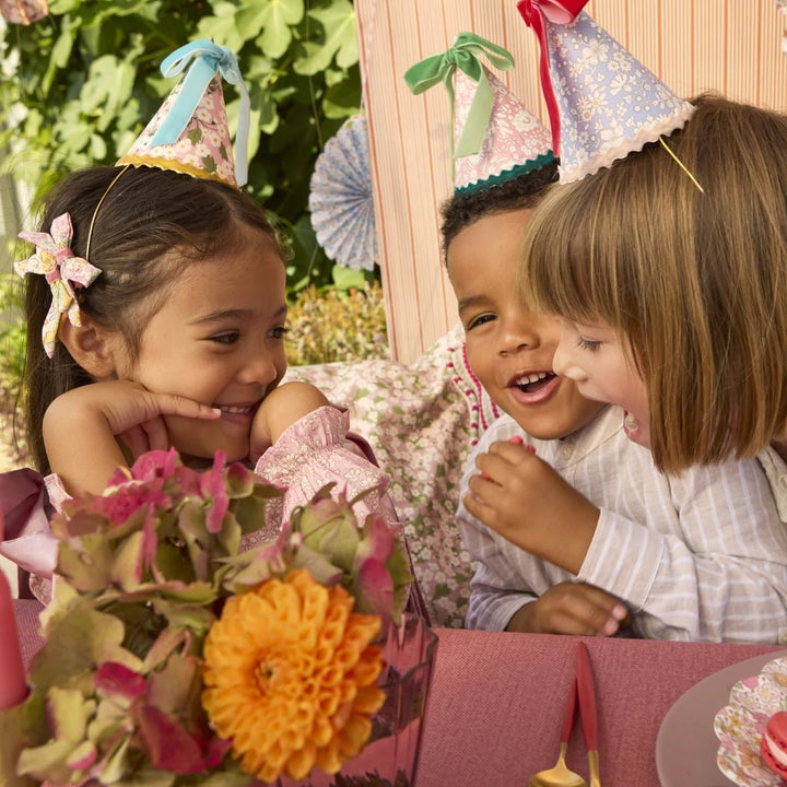 Three children wearing party hats around a table with flowers and a pink tablecloth.