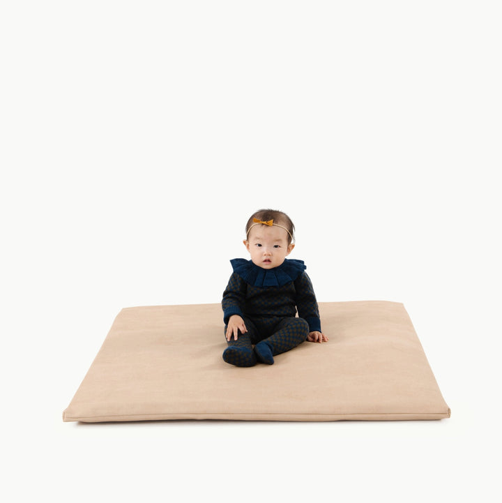 Sand suede padded baby play mat with a baby in a ruffle collar sitting on it.