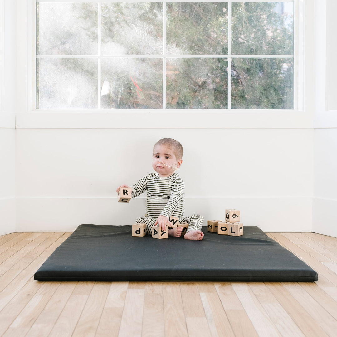 Baby sitting on the black vegan leather padded play mat with wooden alphabet blocks by a window