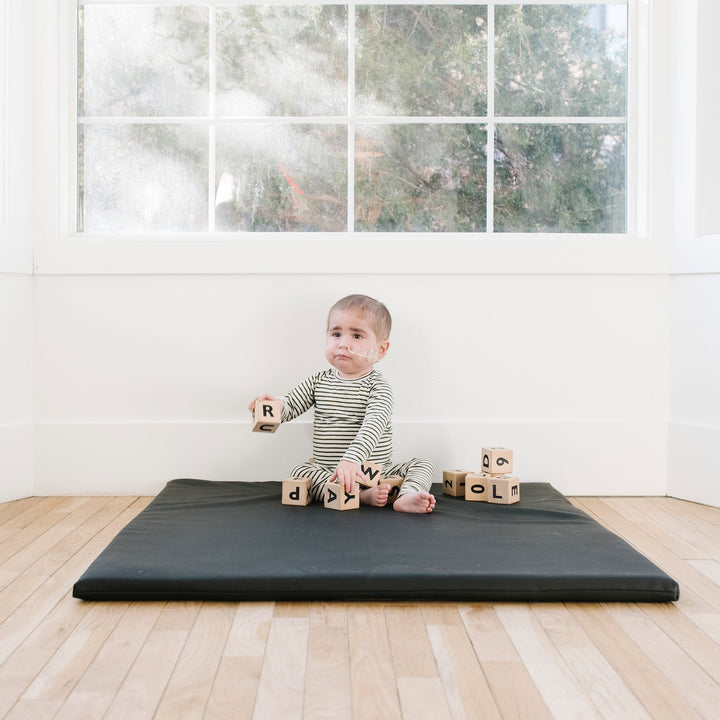 Baby sitting on the black vegan leather padded play mat with wooden alphabet blocks by a window