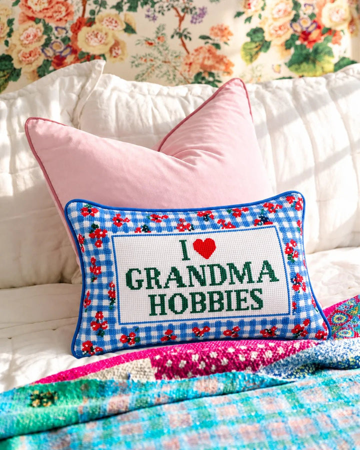 Grandma Hobbies pillow layered on a bed with a pink pillow and floral wallpaper behind