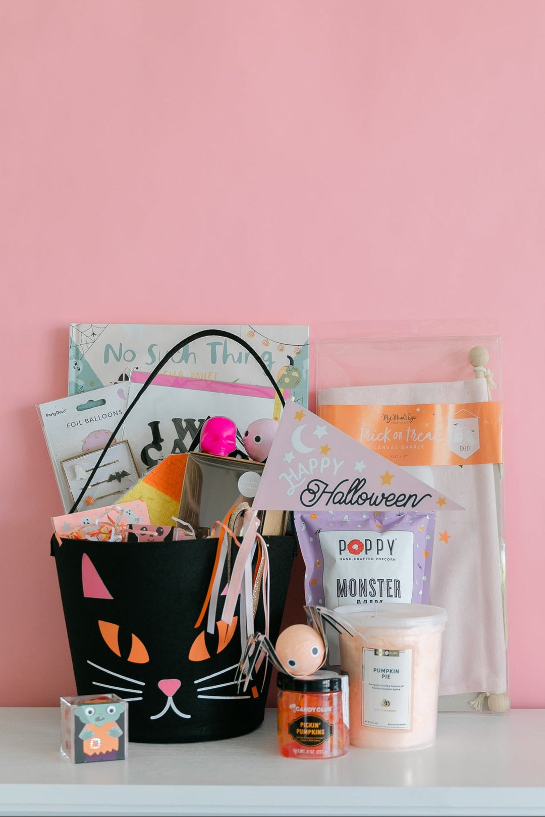 A Halloween-themed gift basket arrangement on a white shelf against a pink wall, featuring a black cat-face bucket filled with treats and cards, alongside a Poppy Monster Candle jar, a small pumpkin, and other seasonal items.