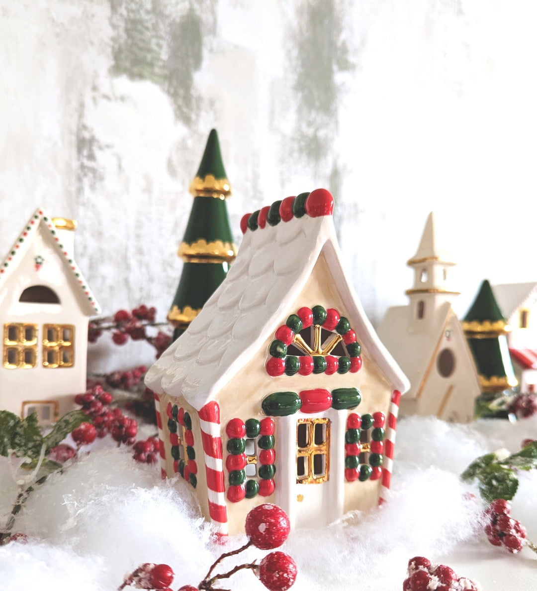 Close-up of a red and green ceramic Christmas cottage among ivory houses, trees, snow, and red berries
