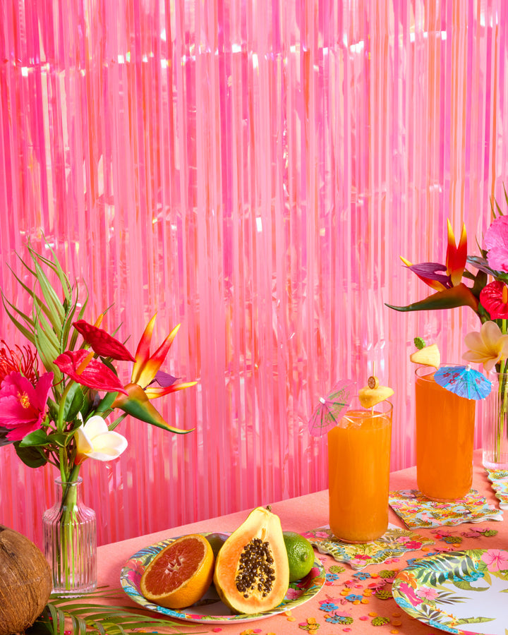 Hot pink iridescent fringe curtain behind a tropical table with papaya, flowers, and drinks