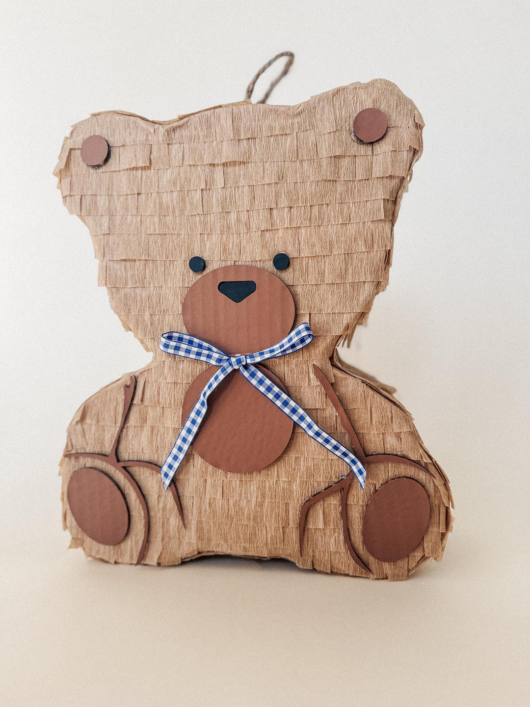 Brown teddy bear-shaped pinata with a blue checkered ribbon on a beige background