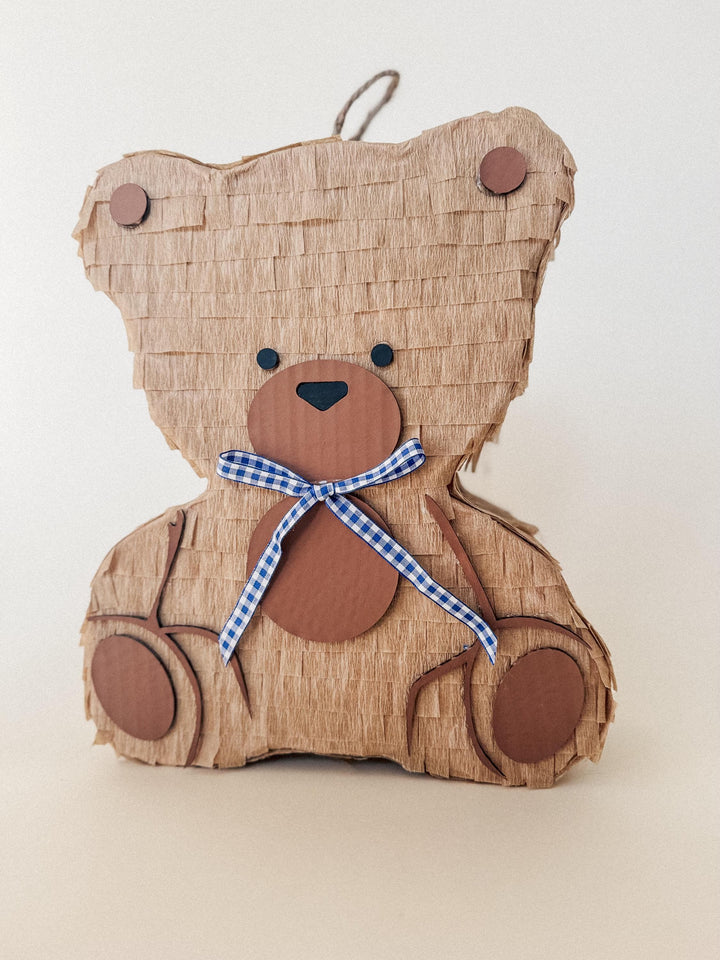 Brown teddy bear-shaped pinata with a blue checkered ribbon on a beige background