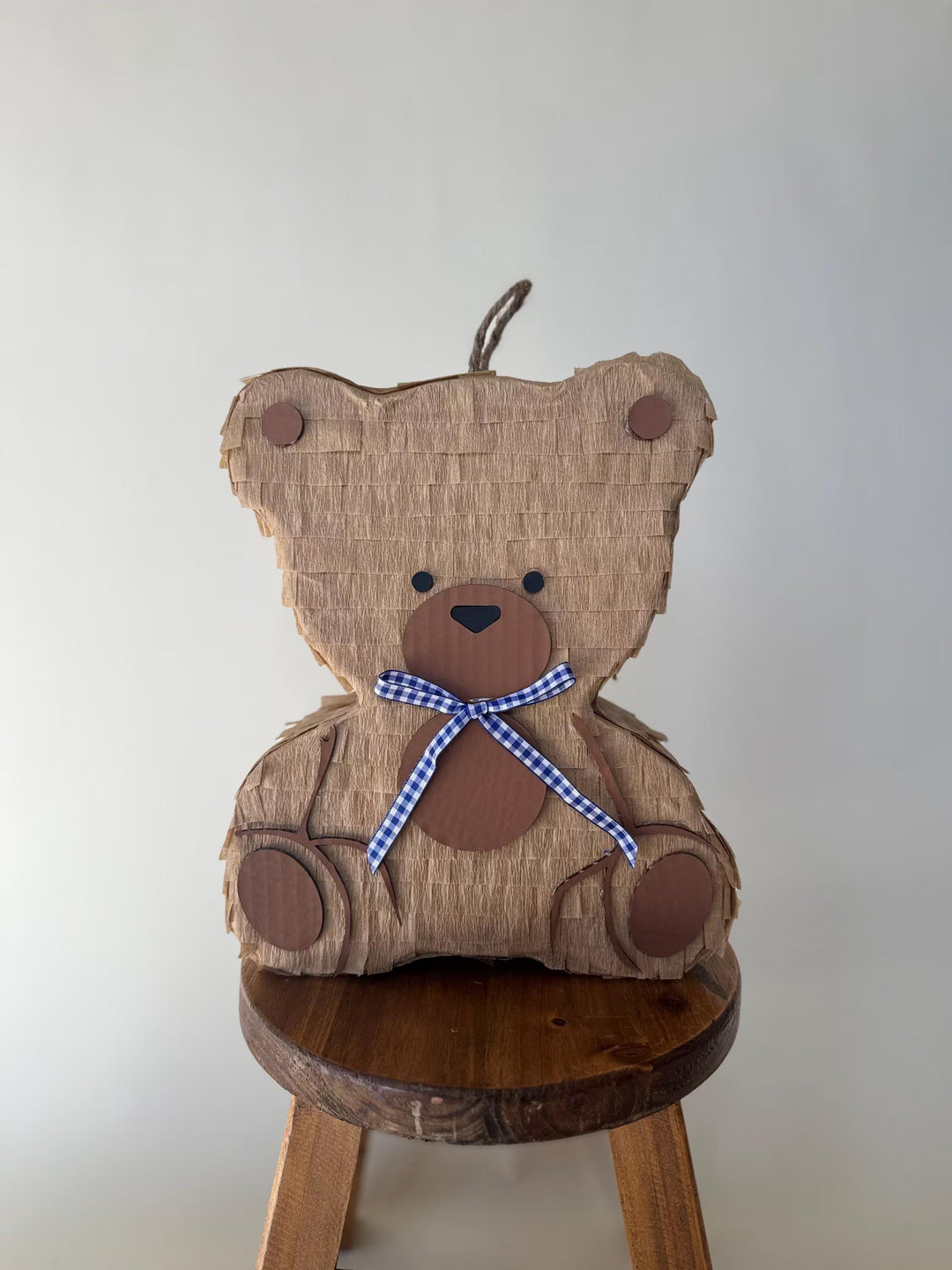 Teddy bear-shaped pinata with a blue ribbon on a wooden stool against a white wall.