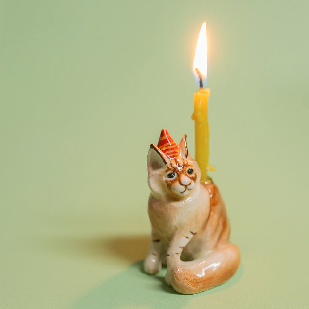 Orange tabby cat birthday candle holder in a striped party hat with a lit candle for a pet birthday cake