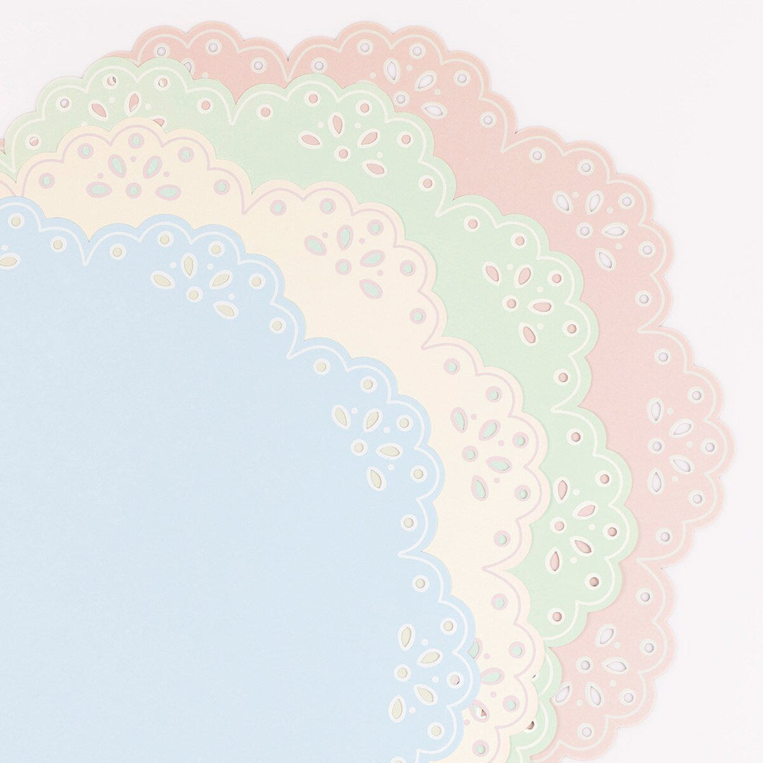 Layered blue, ivory, green and pink paper placemats showing scalloped floral eyelet-style borders.