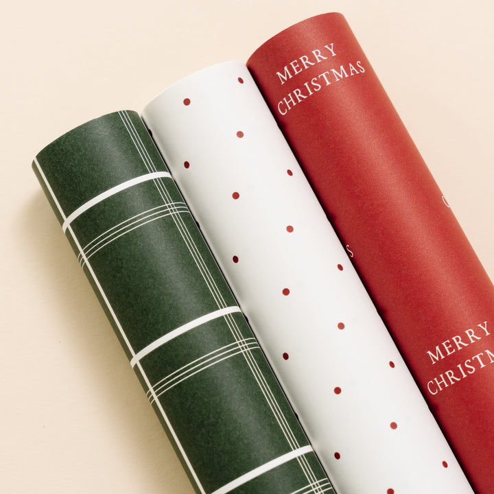 Rolls of green plaid, red polka dot on white, and red Merry Christmas wrapping paper side by side