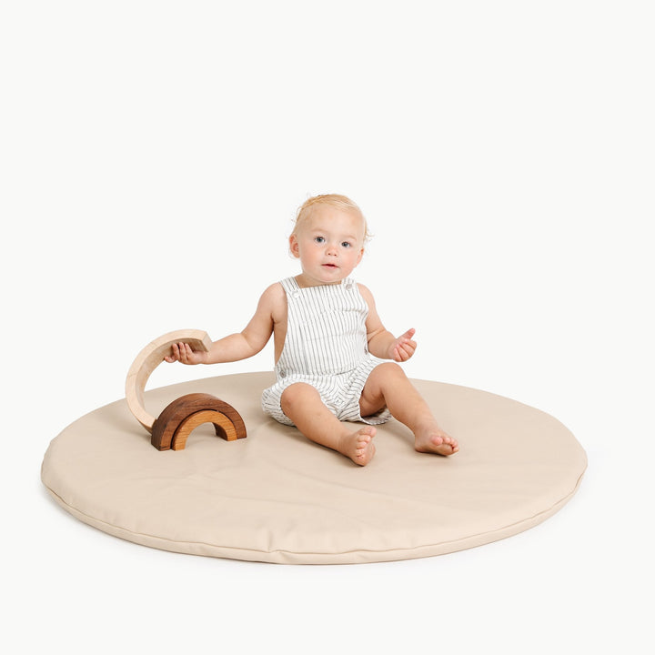 Baby sitting on the round Millet oatmeal padded play mat with a wooden rainbow stacker