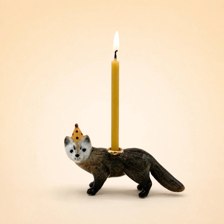 Dark brown porcelain mink cake topper in a tiny party hat holding a lit beeswax candle