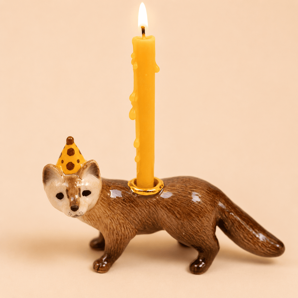 Porcelain mink cake topper with a gold party hat and a lit yellow taper candle, side view