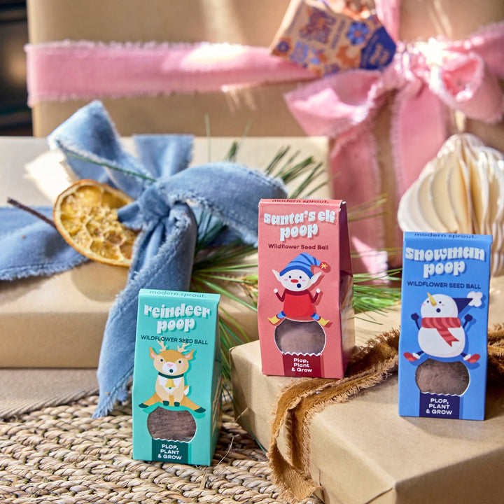 Reindeer, snowman, and Santa's elf poop seed bomb boxes set among kraft-wrapped gifts with pink and blue ribbon