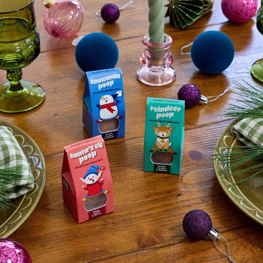 Seed bomb boxes on a wood Christmas dinner table with green goblets, blue ornaments, and a pink candlestick