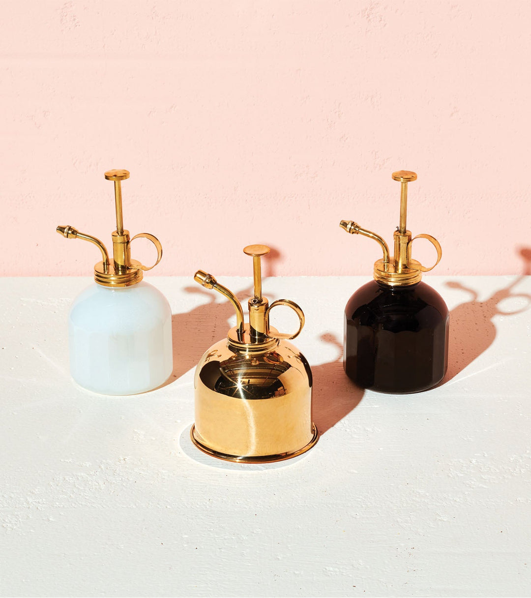 Three plant misters in white, polished brass, and black, each with a brass pump and curved nozzle, on a blush backdrop