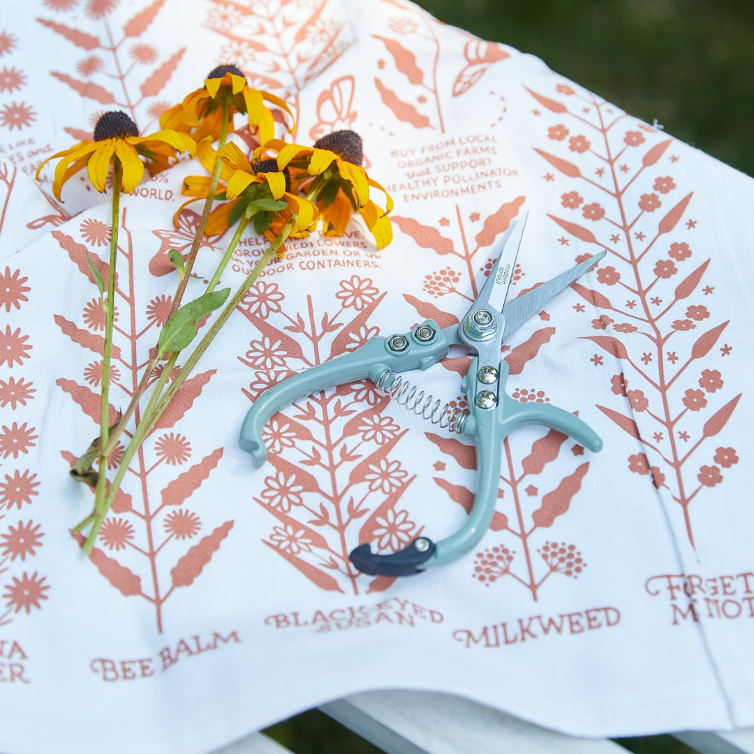 Sage needle-nose shears on a printed wildflower tea towel beside cut yellow black-eyed Susans