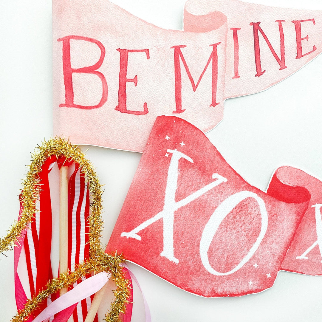 Pink Be Mine and red XOXO party pennants displayed together with striped ribbons and gold tinsel