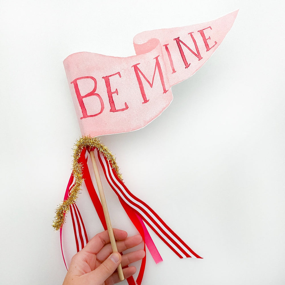 Hand holding a pink Be Mine pennant flag with red-striped and pink ribbons and gold tinsel on a wood dowel
