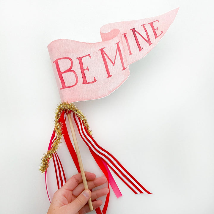 Hand holding a pink Be Mine pennant flag with red-striped and pink ribbons and gold tinsel on a wood dowel