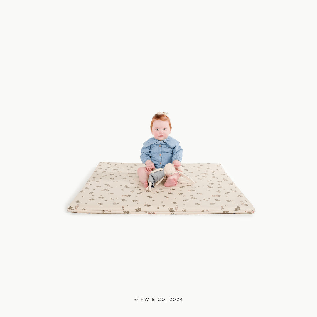 Baby sitting on the square Peter Rabbit padded play mat with a bunny plush