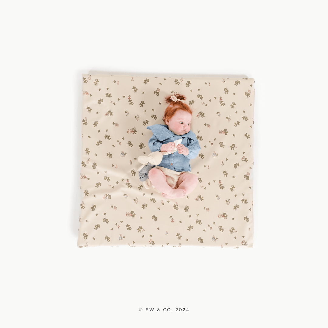 Overhead view of a baby lying on the square Peter Rabbit padded play mat with a bunny plush