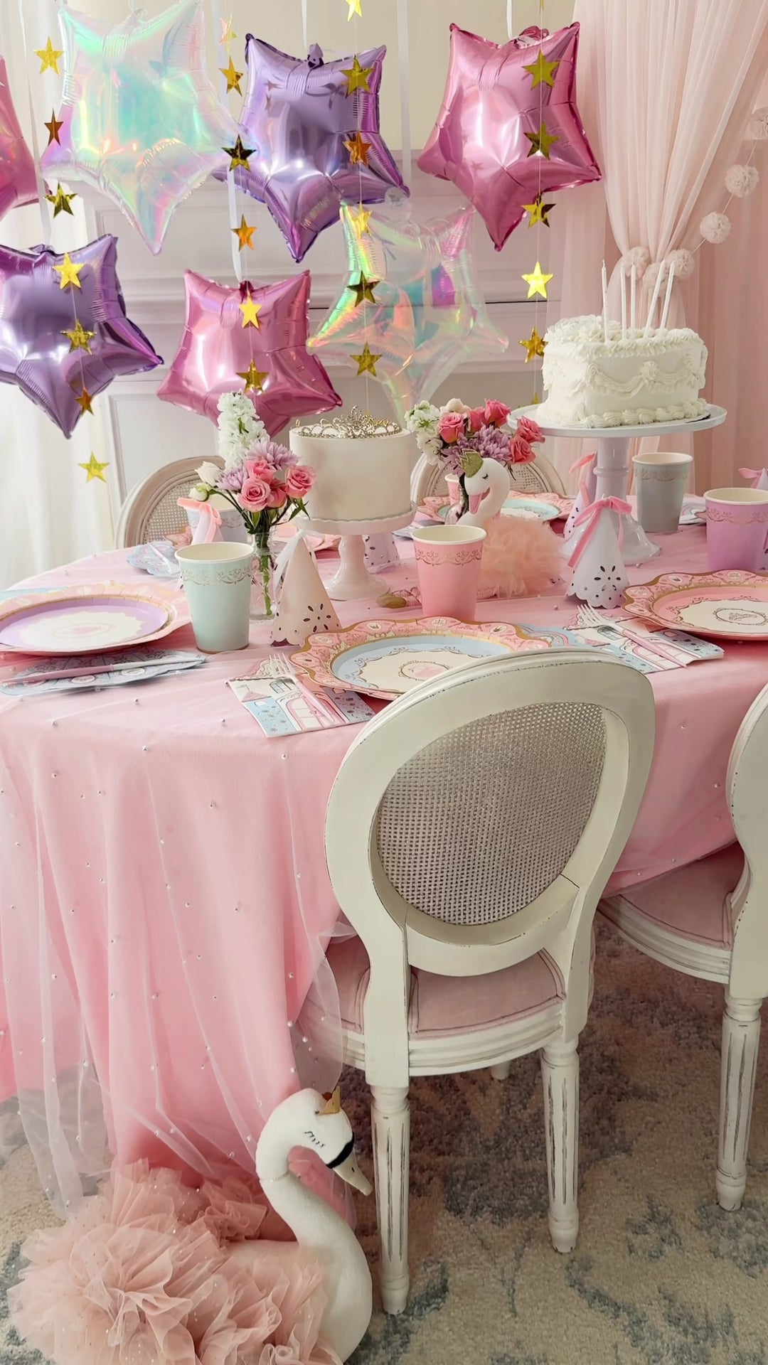 Pastel birthday tablescape with iridescent, lavender, and pink star balloons, gold star garland, a white tiered cake with candles, pink tablecloth, pastel plates and cups, and a white swan accent