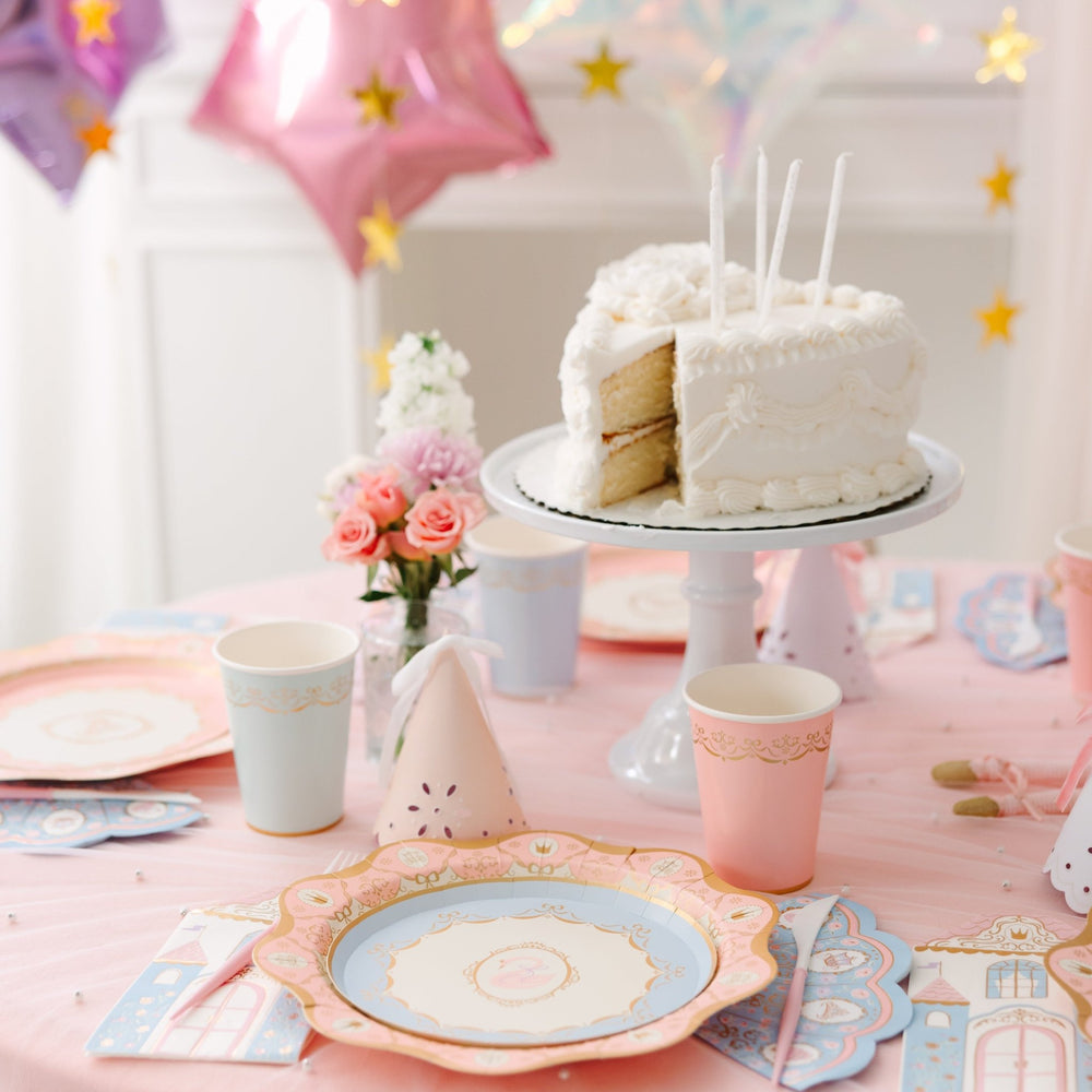 White frosted birthday cake with a slice cut out on a cake stand, surrounded by pink roses, pastel cups, a party hat, and princess plates under star balloons