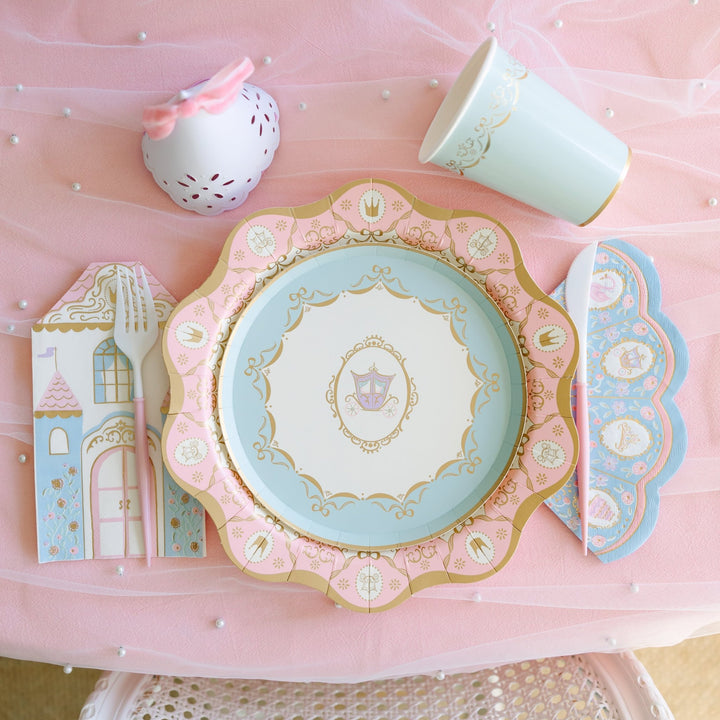 Princess party place setting with a light blue plate featuring a gold crown design on a pink scalloped charger, castle napkin, blue cup, and pink-handled flatware on pink tulle