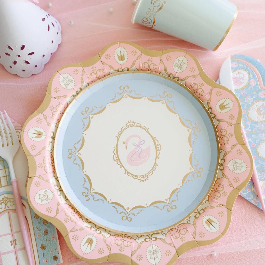 Light blue paper plate with a pink swan illustration in a gold frame, set on a pink scalloped plate with gold crown details next to a gold-rimmed blue cup