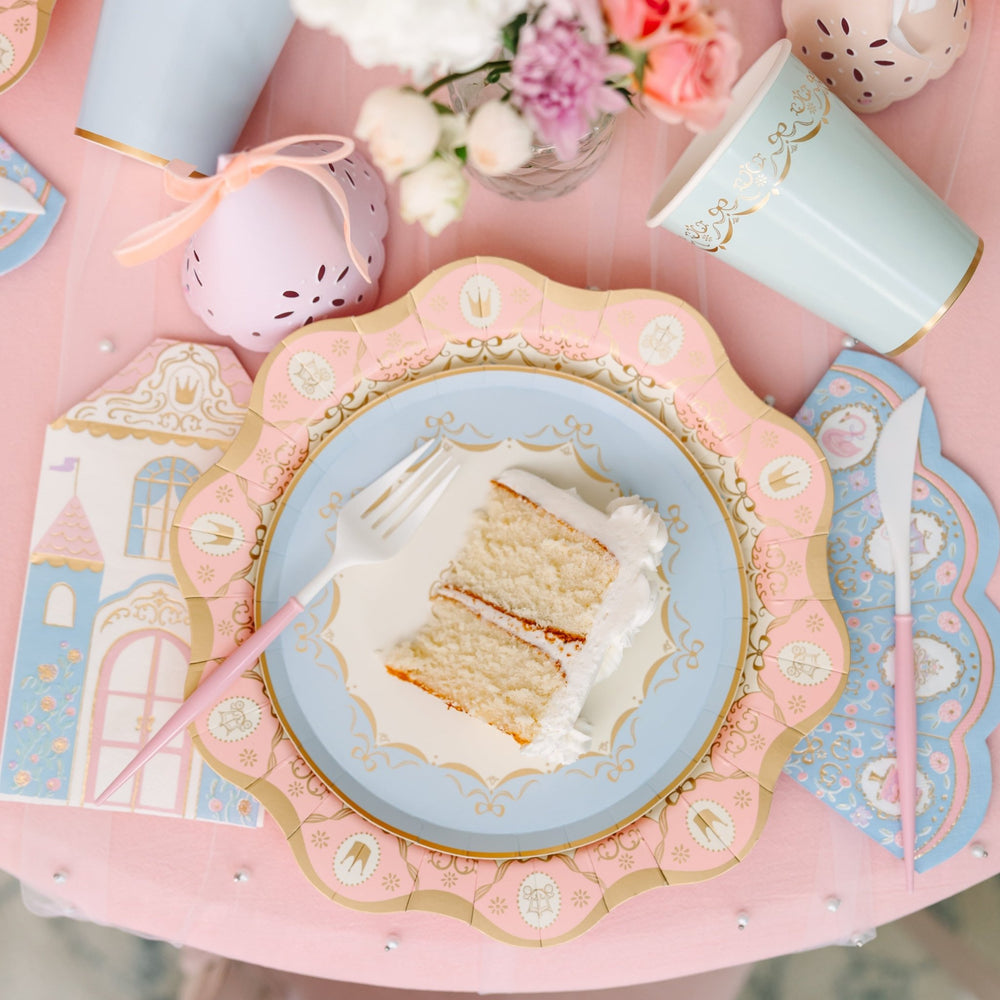 Slice of vanilla layer cake on a scalloped pink and blue princess plate with a castle-shaped napkin, pink-handled fork and knife, and a blue gold-trimmed cup