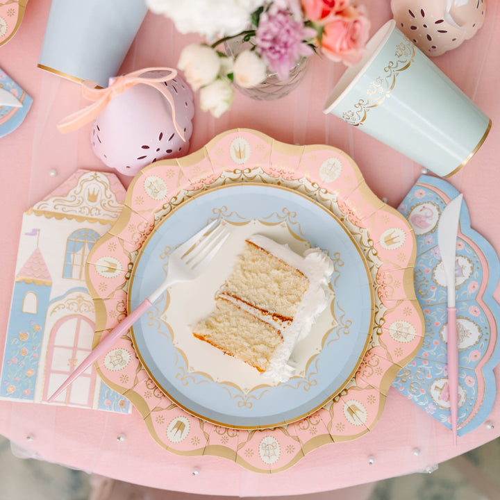 Slice of vanilla layer cake on a scalloped pink and blue princess plate with a castle-shaped napkin, pink-handled fork and knife, and a blue gold-trimmed cup