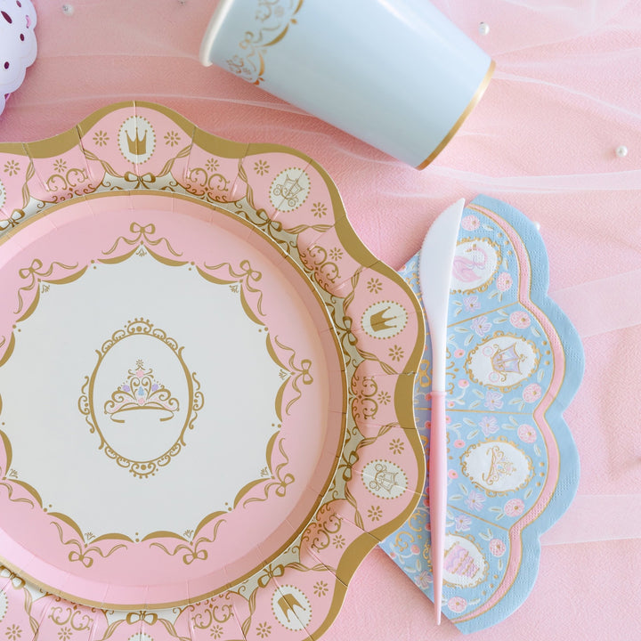 Close-up of a pink scalloped paper plate with gold foil tiara and crown details beside a blue floral scalloped napkin, pink-handled knife, and gold-rimmed blue cup