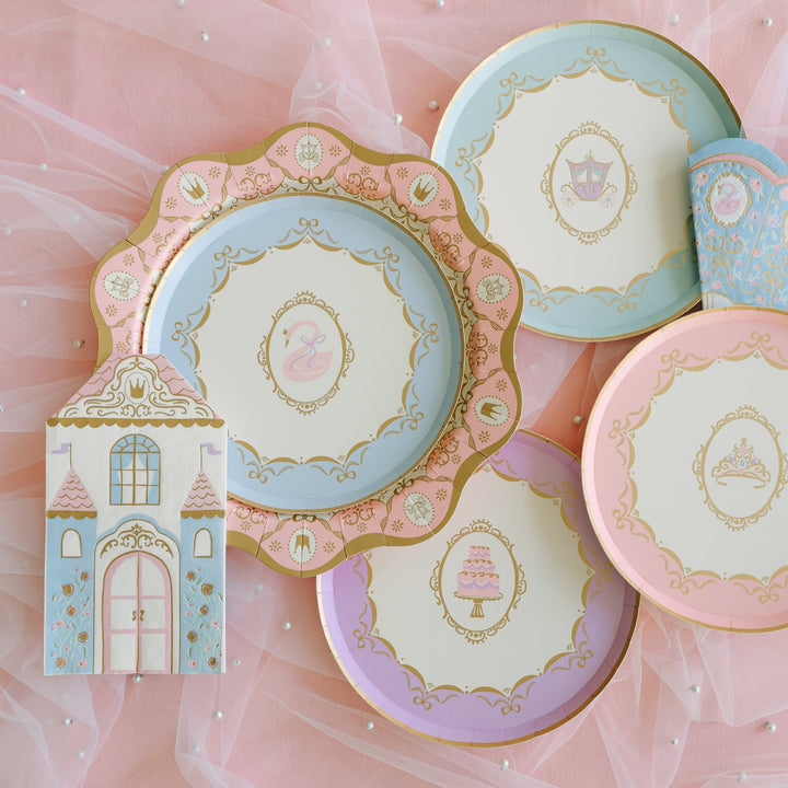 Flat lay of princess party plates in blue, pink, and lavender with gold swan, carriage, tiara, and cake designs, a pink scalloped plate, and a castle-shaped napkin on pink tulle