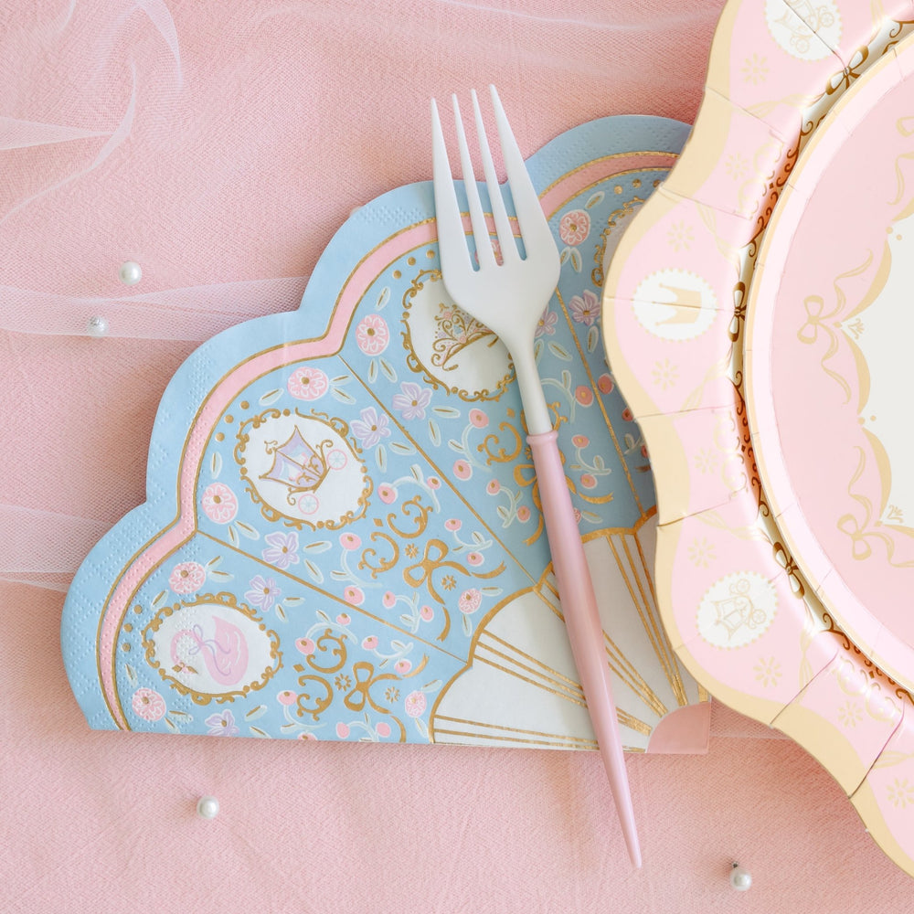 Blue scalloped princess napkin with pink and gold floral and swan details, topped with a pink-handled fork beside a pink scalloped plate on a pink tablecloth with pearls