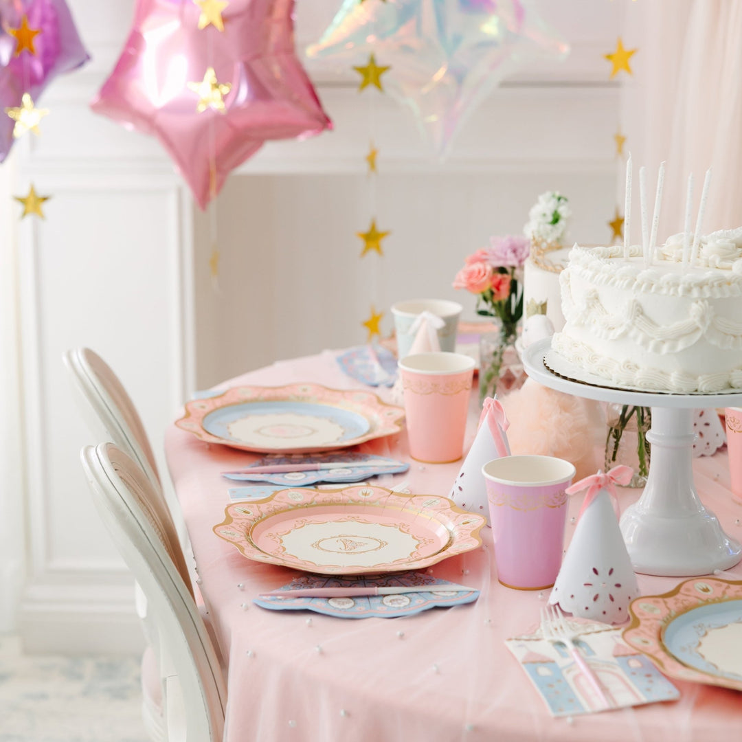 Princess party place settings with scalloped pink and blue plates, pink cups, party hats, and a white birthday cake under pink and iridescent star balloons