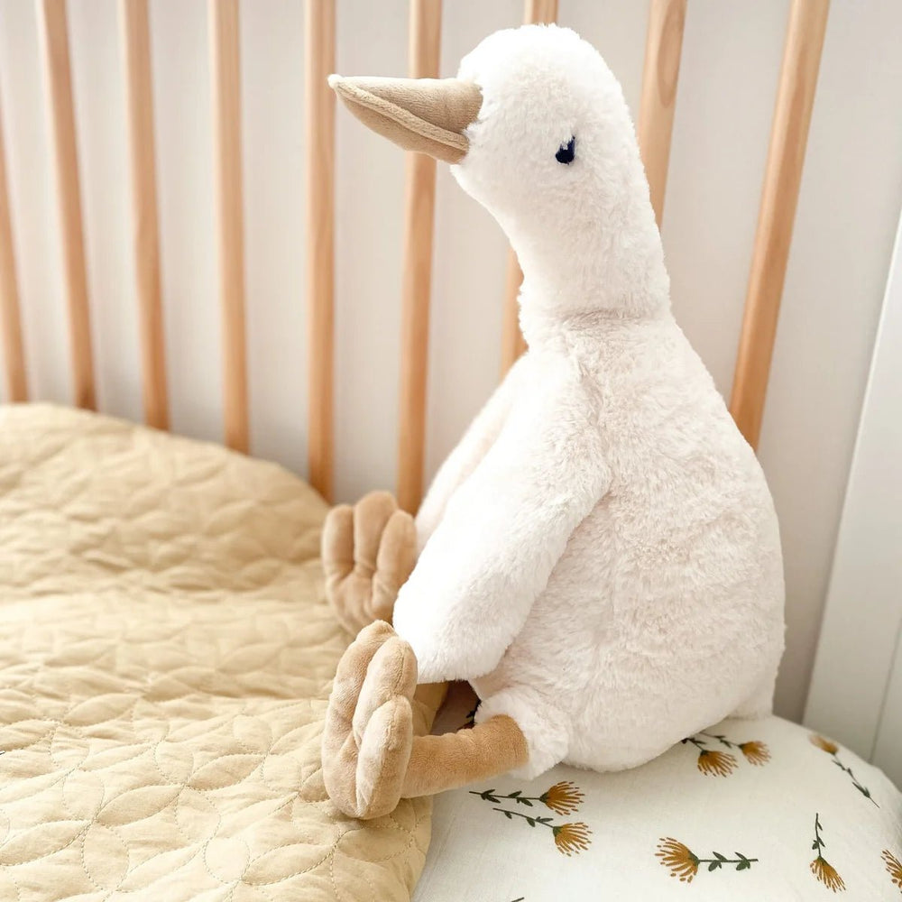 Plush toy goose sitting on a floral pillow with a crib in the background
