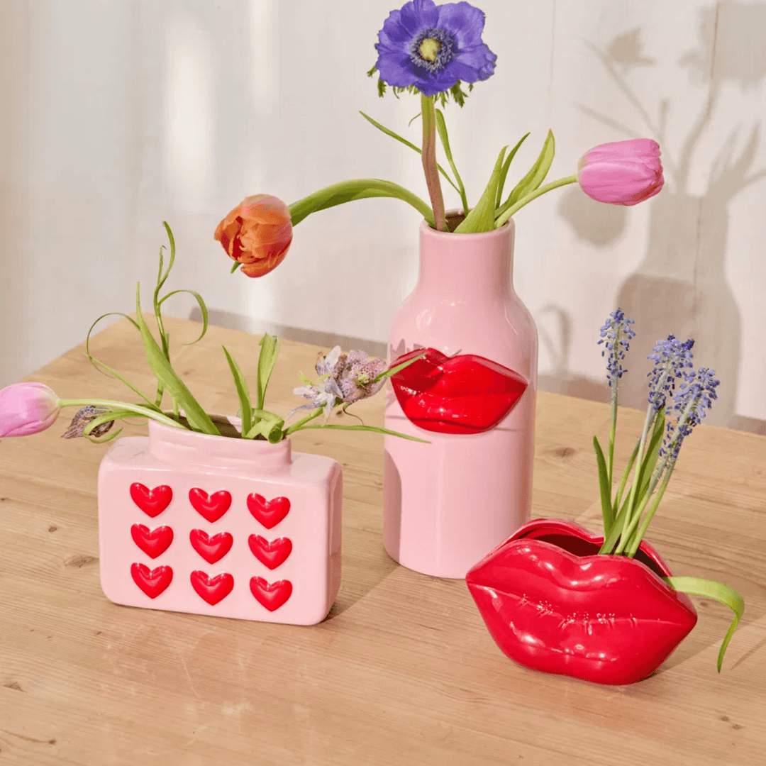 Pink heart vase with spring flowers beside a tall pink bottle vase with red lips and a red lips-shaped vase on a wood table