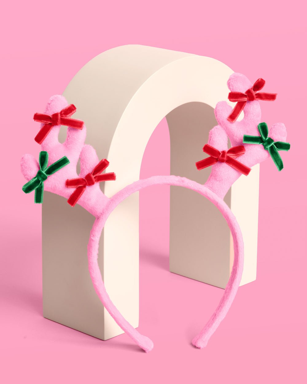 Pink plush reindeer headband standing on a cream arch, showing both bow-tied antlers