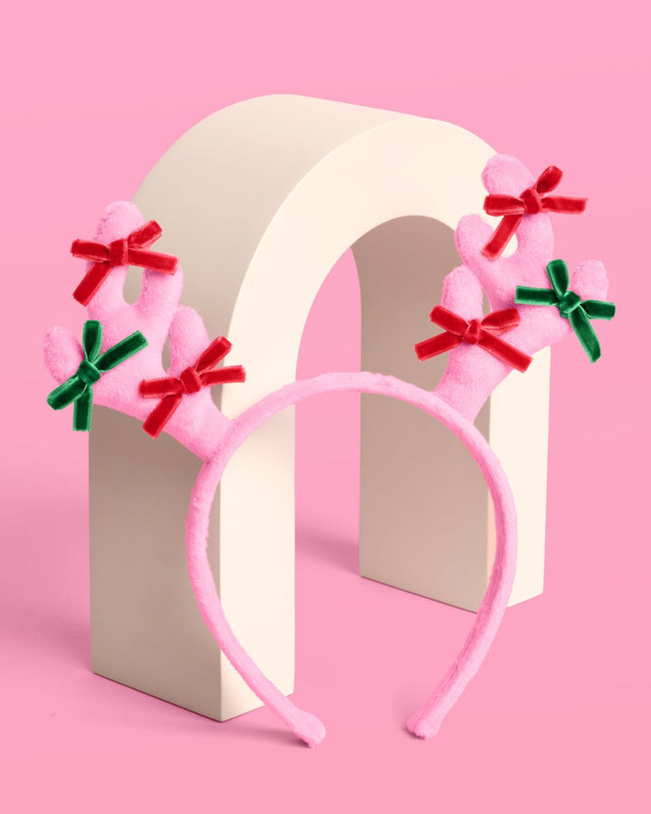 Pink plush reindeer headband standing on a cream arch, showing both bow-tied antlers